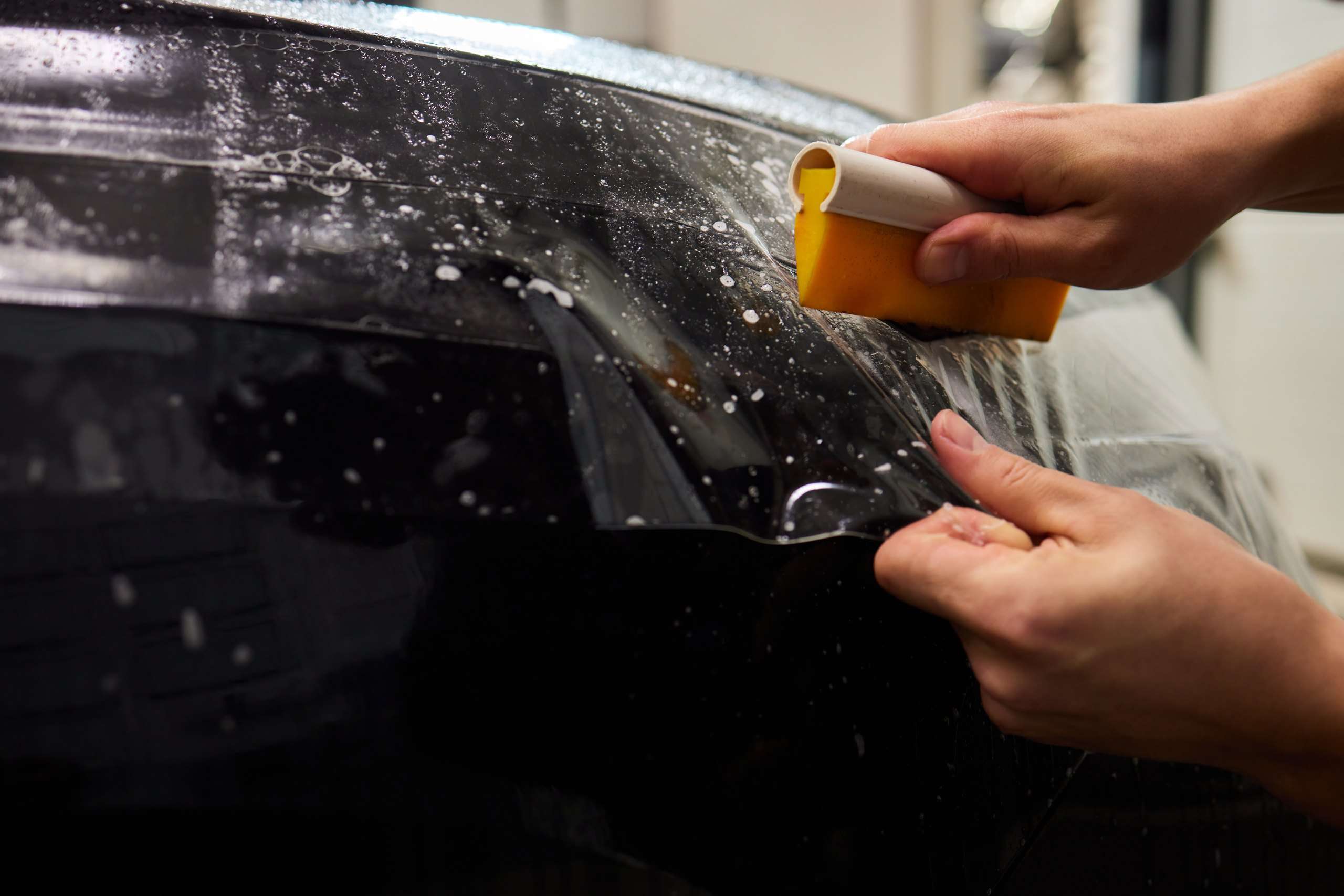 Transparente Lackschutzfolie wird mit einem Rakel nass auf die Front eines schwarzen Autos aufgebracht